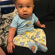Karmelo is registered to the contest to win money with this photo: baby, child, curly_hair, blue_shirt, pajama_pants, cookie_monster, sitting, couch, blanket, indoor, wide_eyes, curious, surprised, baby_bottle, cozy, infant, person, clothing, footwear, home
