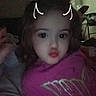child, toddler, portrait, indoors, sitting, pink_shirt, curly_hair, dark_hair, facial_expression, pursed_lips, doodle, horns, bedroom, couch, blanket, arm, close_up, low_light, grainy, family