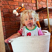 Millie is registered to the contest to win money with this photo: toddler, child, curly_hair, messy_face, basket, white_basket, plush_toy, stuffed_animal, horse_toy, brick_wall, wooden_floor, indoor, casual_clothing, red_stripes, green_trim, necklace, playful, messy, cute, portrait