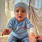Santiano participe au concours pour gagner de l'argent avec cette photo : baby, child, blue_shirt, blue_beanie, jeans, shoe, floor, curtain, indoor, cute, sitting, hand, footwear, light, texture, person, portrait, young_child, casual_clothing, expression