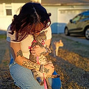 Cookie is registered to the contest to win money with this photo: person, cat, tattoo, grass, harness, outdoor, sunlight, house, car, animal, pet, kneeling, pink, jeans, short_sleeve, tabby_cat, tattooed_arms, daytime, background_cat, nature