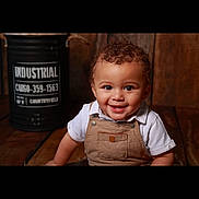 Lucas a rejoint le concours — aidez-le/la à gagner de superbes lots ! toddler, child, smiling, curly_hair, overalls, white_shirt, wooden_floor, indoor, barrel, industrial, portrait, happy, cute, person, baby, sitting, face, eyes, background, rustic
