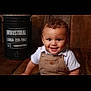 toddler, child, smiling, curly_hair, overalls, white_shirt, wooden_floor, indoor, barrel, industrial, portrait, happy, cute, person, baby, sitting, face, eyes, background, rustic