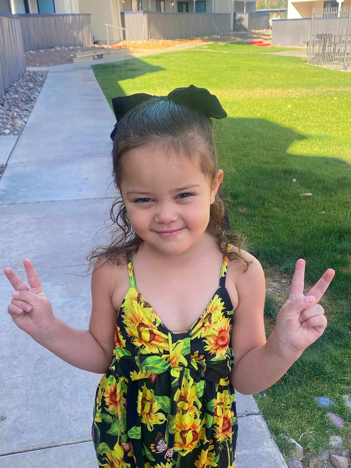 Aleyah is registered to the contest to win money with this photo: child, girl, smiling, peace_sign, sunflower_dress, black_bow, outdoor, sidewalk, grass, residential_area, happy, person, curly_hair, summer, casual_clothing, nature, portrait, young, cute, daylight