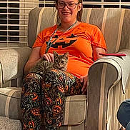Lily joined the competition — help win amazing prizes! woman, cat, armchair, halloween, pajamas, orange_shirt, striped_furniture, indoor, petting, glasses, smiling, tabby_cat, phone, window_blinds, casual, sitting, comfortable, happy, cozy, home