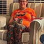 woman, cat, armchair, halloween, pajamas, orange_shirt, striped_furniture, indoor, petting, glasses, smiling, tabby_cat, phone, window_blinds, casual, sitting, comfortable, happy, cozy, home