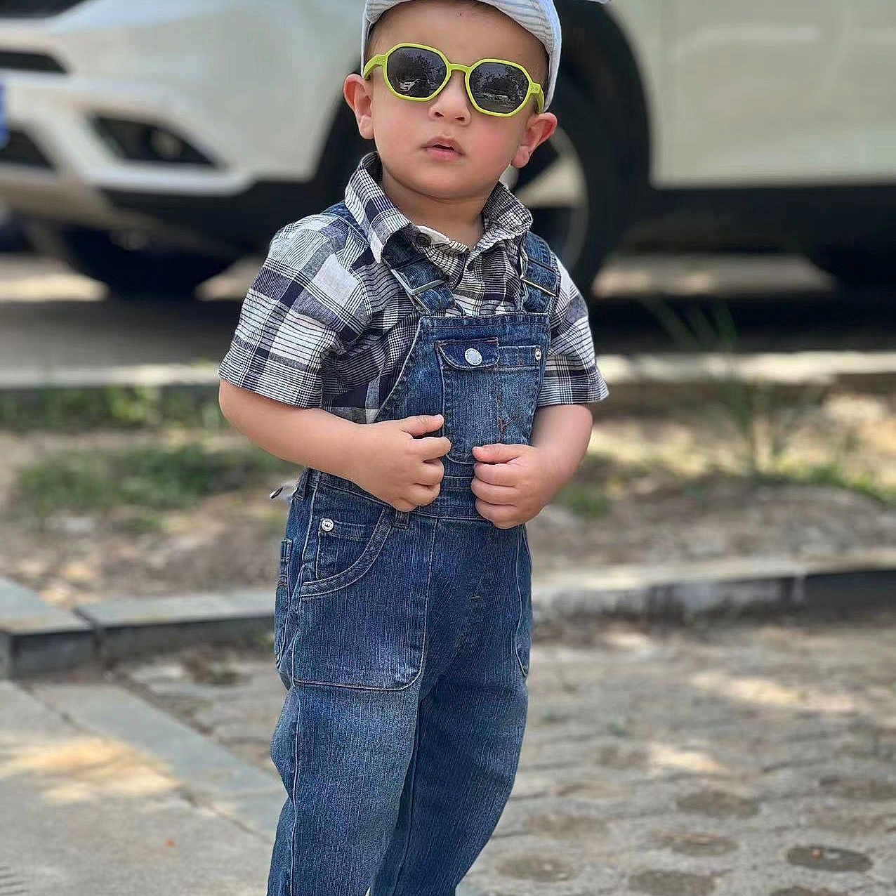 Aliyan is registered to the contest to win money with this photo: accessory, bunny_ears, car, casual_clothing, child, cute, denim_overalls, fashion, footwear, outdoor, pavement, person, plaid_shirt, playful, sidewalk, standing, sunglasses, sunny, toddler, young_child