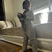 Aissata participe au concours pour gagner de l'argent avec cette photo : child, toddler, indoor, standing, toy, soft_toy, window, natural_light, hair_beads, socks, pajamas, carpet, couch, cushions, floor, radiator, curtains, home, person, portrait