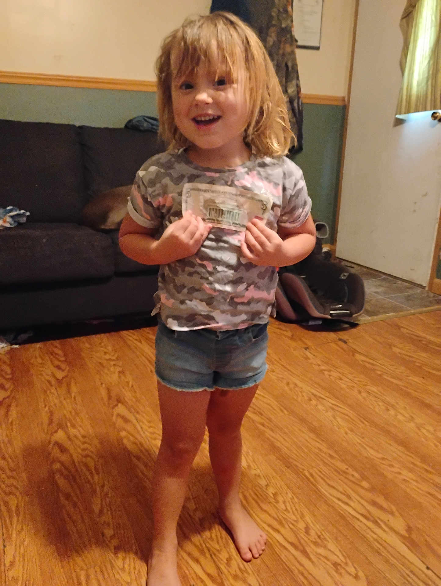 Penelope is registered to the contest to win money with this photo: child, barefoot, smiling, indoor, wooden_floor, denim_shorts, camo_shirt, blond_hair, living_room, sofa, door, curtains, carpet, happy, person, casual_clothing, home, flooring, standing, young