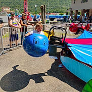 Penelope is registered to the contest to win money with this photo: child, carnival_ride, blue_whale, outdoor, sunny, people, fence, background, hill, building, picnic_table, casual_clothing, daytime, amusement_park, ride_seat, shadow, curious_expression, spectators, hat, glasses