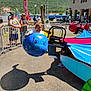 child, carnival_ride, blue_whale, outdoor, sunny, people, fence, background, hill, building, picnic_table, casual_clothing, daytime, amusement_park, ride_seat, shadow, curious_expression, spectators, hat, glasses