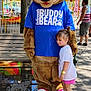 Penelope joined the competition — help win amazing prizes! child, mascot, bear_costume, amusement_park, outdoor, girl, pink_shorts, purple_sneakers, blue_shirt, cap, bench, tree, pavement, sunlight, people, fence, ride, daytime, fun, hug