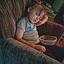 child, sleeping, armchair, plastic_container, indoor, casual_clothing, toddler, relaxed, home, furniture, young_child, resting, peaceful, curly_hair, closed_eyes, seated, dim_lighting, carpet, floor, cozy