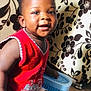 baby, blue_object, child, cute, face, floor, hand, happy, home, indoor, knitted_clothing, patterned_cushion, person, portrait, red_vest, short_hair, sitting, skin, smiling, toddler