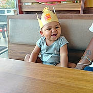 Maëlan a rejoint le concours — aidez-le/la à gagner de superbes lots ! toddler, child, burger_king, crown, smile, happy, restaurant, booth, wood, cushion, window, daylight, casual_clothing, person, indoor, portrait, cute, seated, fun, cheerful