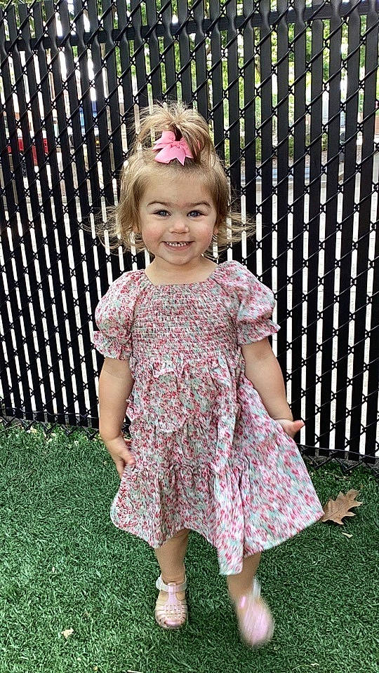 Mariella is registered to the contest to win money with this photo: baby_toddler_clothing, clothing, day_dress, dress, face, fence, fun, grass, happy, head, joy, magenta, one_piece_garment, pattern, person, pink, purple, sleeve, smile, summer