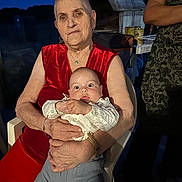 Isaël participe au concours pour gagner de l'argent avec cette photo : baby, blue_pants, bracelet, casual, chair, elderly_woman, expression, family, gathering, holding, lighting, necklace, night, outdoor, person, portrait, red_clothing, seated, watch, white_shirt