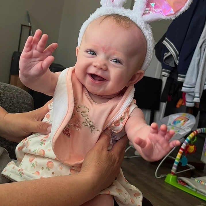 Symphanie is registered to the contest to win money with this photo: adult_hand, baby, bib, bunny_ears, carpet, child, clothing, cute, floor, furniture, hands, happy, home, indoor, infant, person, playful, portrait, smiling, toy