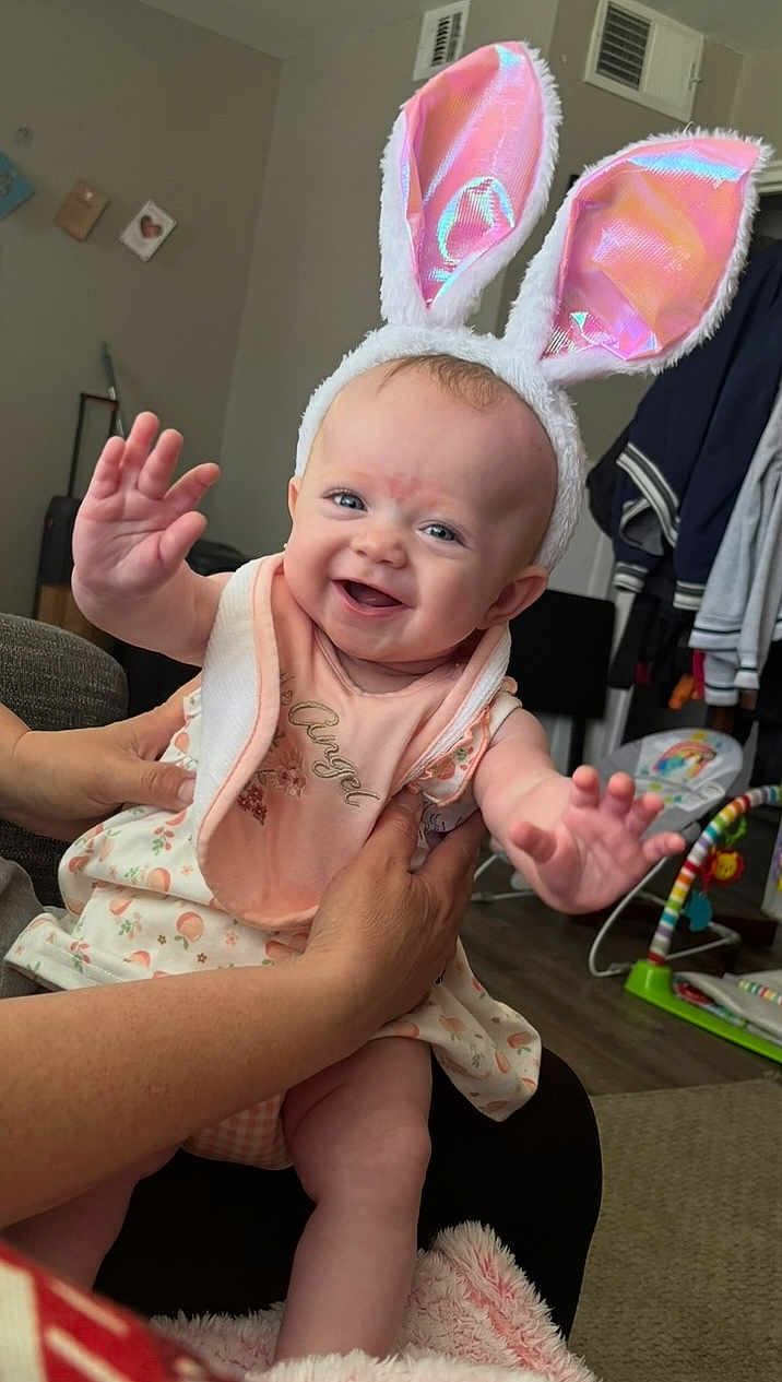 Symphanie is registered to the contest to win money with this photo: baby, bunny_ears, smiling, child, indoor, person, hands, clothing, bib, playful, happy, home, furniture, carpet, toy, floor, adult_hand, cute, infant, portrait