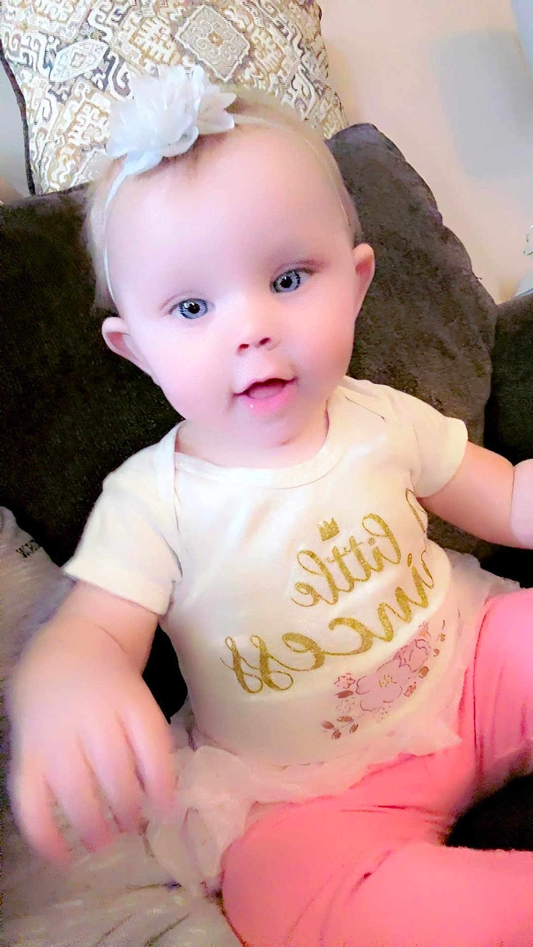Mia is registered to the contest to win money with this photo: baby, child, infant, face, smile, headband, flower, blue_eyes, couch, pillow, clothing, white_shirt, pink_pants, toddler, sitting, cute, indoors, person, happy, portrait
