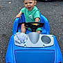 toddler, child, toy_car, blue, green_shirt, steering_wheel, outdoor, asphalt, happy, smiling, short_hair, sitting, play, vehicle, plastic, wheel, daylight, expression, cute, small