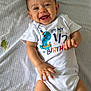 baby, infant, smiling, happy, onesie, white_clothing, text_on_clothing, birthday, lying_down, blanket, patterned_blanket, cute, child, face, hands, skin, indoors, young_child, celebration, portrait