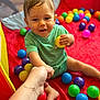 toddler, child, baby, green_shirt, red_mat, colorful_balls, yellow_ball, hand_holding, indoor, play_area, smiling, happy, person, face, skin, foot, arm, toy, playtime, casual
