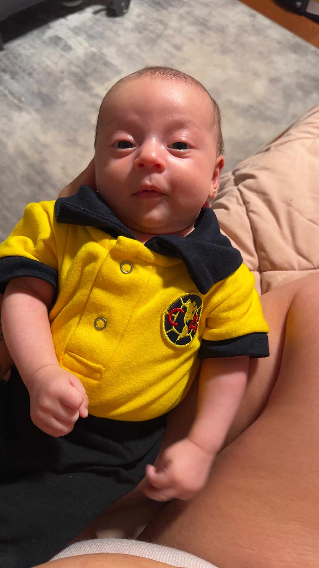 Ailani joined the competition — help win amazing prizes! baby, infant, yellow_shirt, black_collar, person, face, hand, lap, skin, indoor, carpet, furniture, closeup, cute, child, young, expression, casual_clothing, sitting, resting