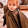 toddler, child, haircut, cape, chair, washing_machine, laundry_room, face, eyes, hair, expression, indoors, door, mat, clothing, person, portrait, seated, curious, cute