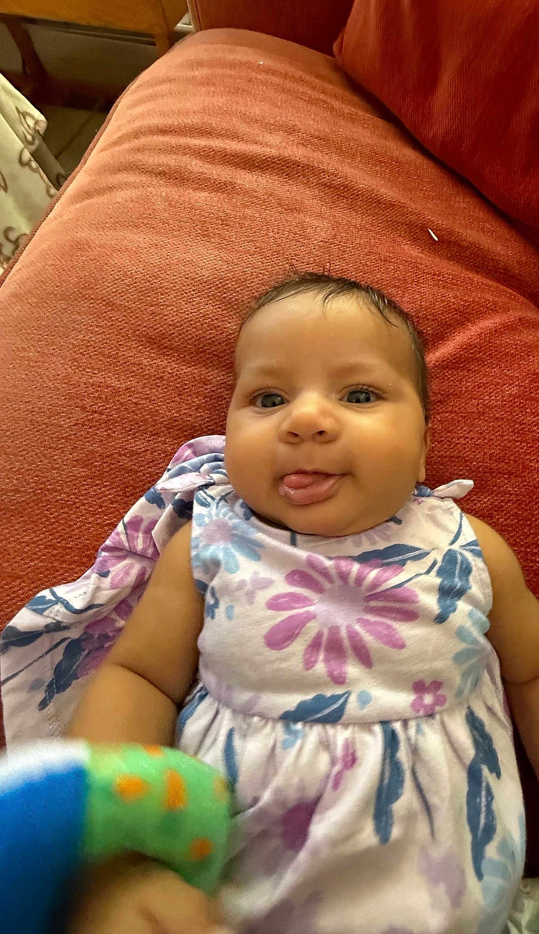 Saphire joined the competition — help win amazing prizes! baby, infant, child, couch, red_couch, floral_dress, tongue_out, toy, colorful_toy, indoor, smiling, happy, cute, face, skin, person, sitting, fabric, home, playful
