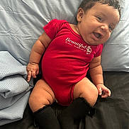 Aurora is registered to the contest to win money with this photo: baby, infant, smiling, red_onesie, black_socks, bed, blanket, blue_blanket, curly_hair, cute, happy, chubby_arms, chubby_legs, person, indoors, portrait, child, resting, soft_lighting, cozy
