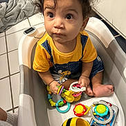 Aurora is registered to the contest to win money with this photo: baby_toys, barefoot, child, colorful, curious, curly_hair, cute, expression, floor_tiles, home, indoor, infant, person, plastic_tub, playing, sitting, toddler, toys, yellow_shirt, young_child