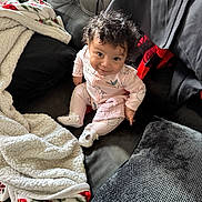Aurora is registered to the contest to win money with this photo: baby, blanket, casual, child, couch, cozy, curly_hair, cute, expression, floor, home, indoor, infant, leather, looking_up, pillow, pink_outfit, sitting, smile, wooden_floor