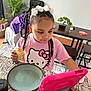 child, girl, braids, white_bows, pink_shirt, hello_kitty, sandwich, tablet, pink_case, kitchen_counter, plate, cup, plant, dining_table, chair, indoor, home, carpet, wood_floor, casual