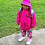child, pink_hat, hoodie, camo_pants, white_sneakers, grass, sidewalk, outdoor, casual_clothing, braided_hair, young_person, smiling, standing, daylight, footwear, fashion, nature, greenery, street, kid