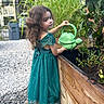 child, girl, teal_dress, watering_can, turtle_shape, garden, plants, greenery, wooden_garden_bed, gravel_path, outdoor, curly_hair, shoes, nature, daylight, portrait, cute, playful, activity, summer