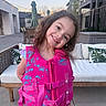 child, girl, smiling, life_jacket, pink, outdoor, patio, cushion, sofa, table, string_lights, umbrella, hair, clothing, person, happy, daytime, building, residential, playful
