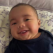 Enzo is registered to the contest to win money with this photo: baby, smiling, tongue_out, face, cheeks, lying_down, pillow, patterned, clothing, navy_blue, cute, infant, head, skin, happy, person, indoors, closeup, portrait, child