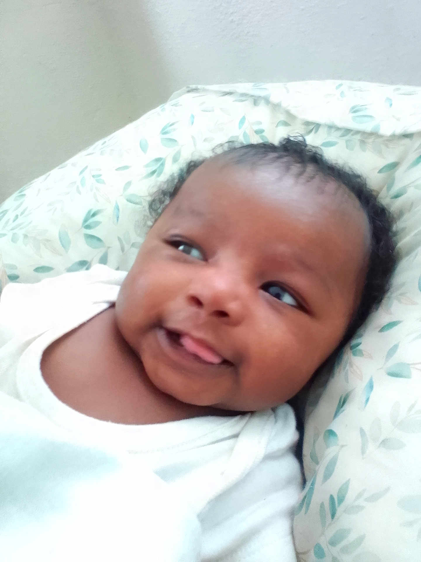 Kadeem is registered to the contest to win money with this photo: baby, infant, face, tongue, smile, curly_hair, pillow, pattern, white_clothing, skin, person, cute, lying_down, indoors, soft_texture, young_child, head, expression, cheeks, playful