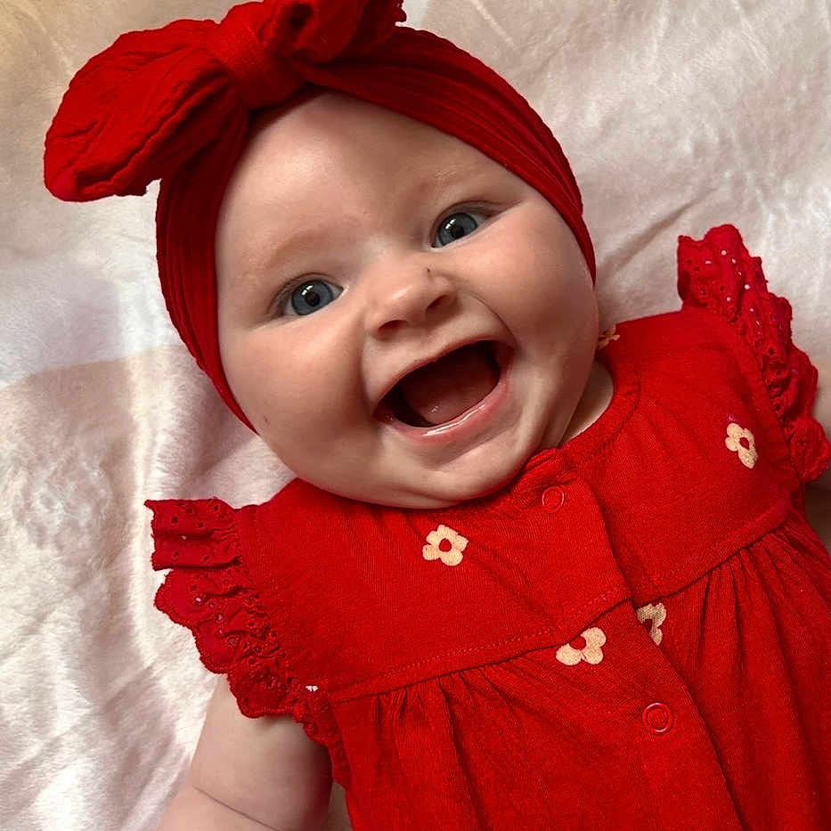 Lilou participe au concours pour gagner de l'argent avec cette photo : apparel, baby, baby_girl, blanket, bow, cheerful, child, closeup, cute, face, flower_pattern, happy, headband, infant, laying, portrait, red_clothing, skin, smiling, young