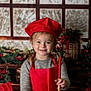 child, smiling, chef_hat, red_apron, wooden_bowl, whisk, spoon, kitchen, christmas_decorations, holiday, snowy_window, baking, flour_bag, festive, gingerbread_card, wooden_table, pigtails, sweater, indoors, happy