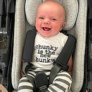 Maximus is registered to the contest to win money with this photo: baby, smiling, infant, stroller, seatbelt, striped_pants, white_shirt, text_on_clothing, happy, cute, child, face, clothing, straps, cushion, infant_clothing, person, sitting, indoor, portrait