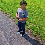 Elliot is registered to the contest to win money with this photo: boy, casual_clothing, child, curly_hair, daylight, grass, greenery, nature, outdoor, pants, park, pavement, portrait, shadow, sidewalk, sneakers, standing, sunny, tshirt, young