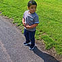 boy, casual_clothing, child, curly_hair, daylight, grass, greenery, nature, outdoor, pants, park, pavement, portrait, shadow, sidewalk, sneakers, standing, sunny, tshirt, young