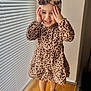toddler, child, leopard_print, dress, headband, barefoot, standing, window_blinds, wood_floor, indoor, smiling, hands_on_face, playful, happy, cute, fashion, portrait, girl, soft_light, home