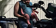 Declan is registered to the contest to win money with this photo: woman, baby, baby_carrier, atv, outdoor, trees, shed, smile, person, vehicle, sunlight, shadow, casual_clothing, nature, greenery, hairstyle, parent, child, daytime, relaxation
