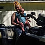 woman, baby, baby_carrier, atv, outdoor, trees, shed, smile, person, vehicle, sunlight, shadow, casual_clothing, nature, greenery, hairstyle, parent, child, daytime, relaxation