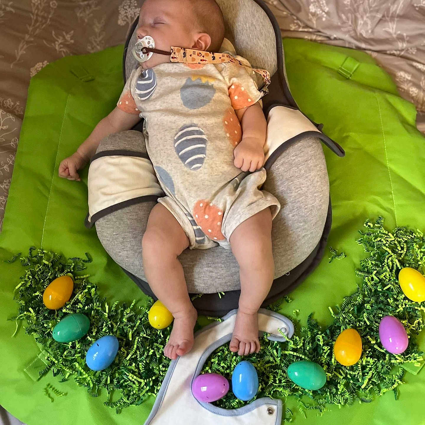 Declan is registered to the contest to win money with this photo: baby, baby_seat, bed, bib, blanket, celebration, child, clothing, cute, easter, easter_eggs, footwear, green_shredded_paper, holiday, indoors, infant, pacifier, person, sleeping, text_on_bib
