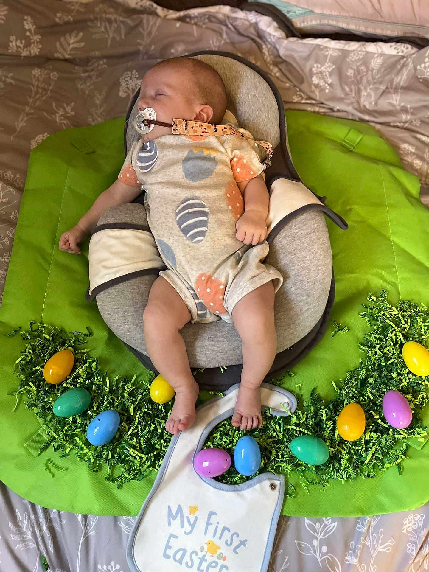 Declan is registered to the contest to win money with this photo: baby, infant, pacifier, sleeping, baby_seat, easter, easter_eggs, green_shredded_paper, bib, text_on_bib, bed, blanket, clothing, footwear, indoors, holiday, cute, child, person, celebration