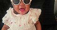 Azure is registered to the contest to win money with this photo: accessory, baby, bow, cheerful, child, closeup, cute, dark_background, fashion, floral_pattern, happy, headband, indoors, infant, person, portrait, sitting, smiling, sunglasses, white_dress